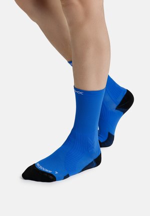 Benen met felblauwe atletieksokken tot halverwege de kuit, met zwarte tenen en hiel, staand op een wit oppervlak.