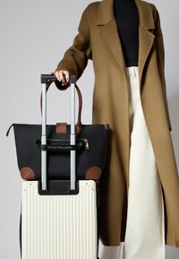 Borsa tote nera con dettagli in pelle marrone, con zip, su una valigia rigida bianca a righe. Modella con cappotto marrone e pantaloni crema.