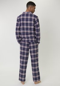 Hombre vestido con pijamas de cuadros en tonos azul marino, blanco y rojo, de pie descalzo mirando hacia atrás con las manos en los bolsillos, contra un fondo claro y liso.