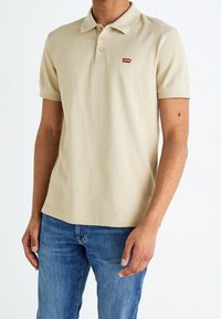 Beige poloko s krátkým rukávem z bavlny. Obsahuje tříknoflíkovou légu a malé červené logo Levi's na levé straně hrudi.