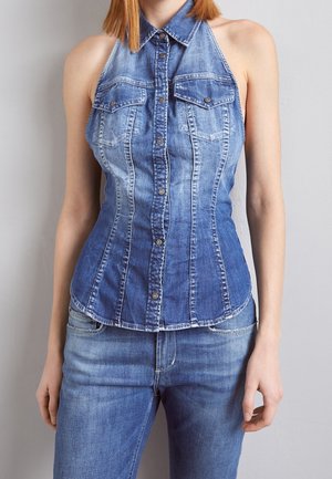 Femme portant un haut en jean bleu sans manches avec col et poches devant, assorti à un jean bleu sur fond gris.