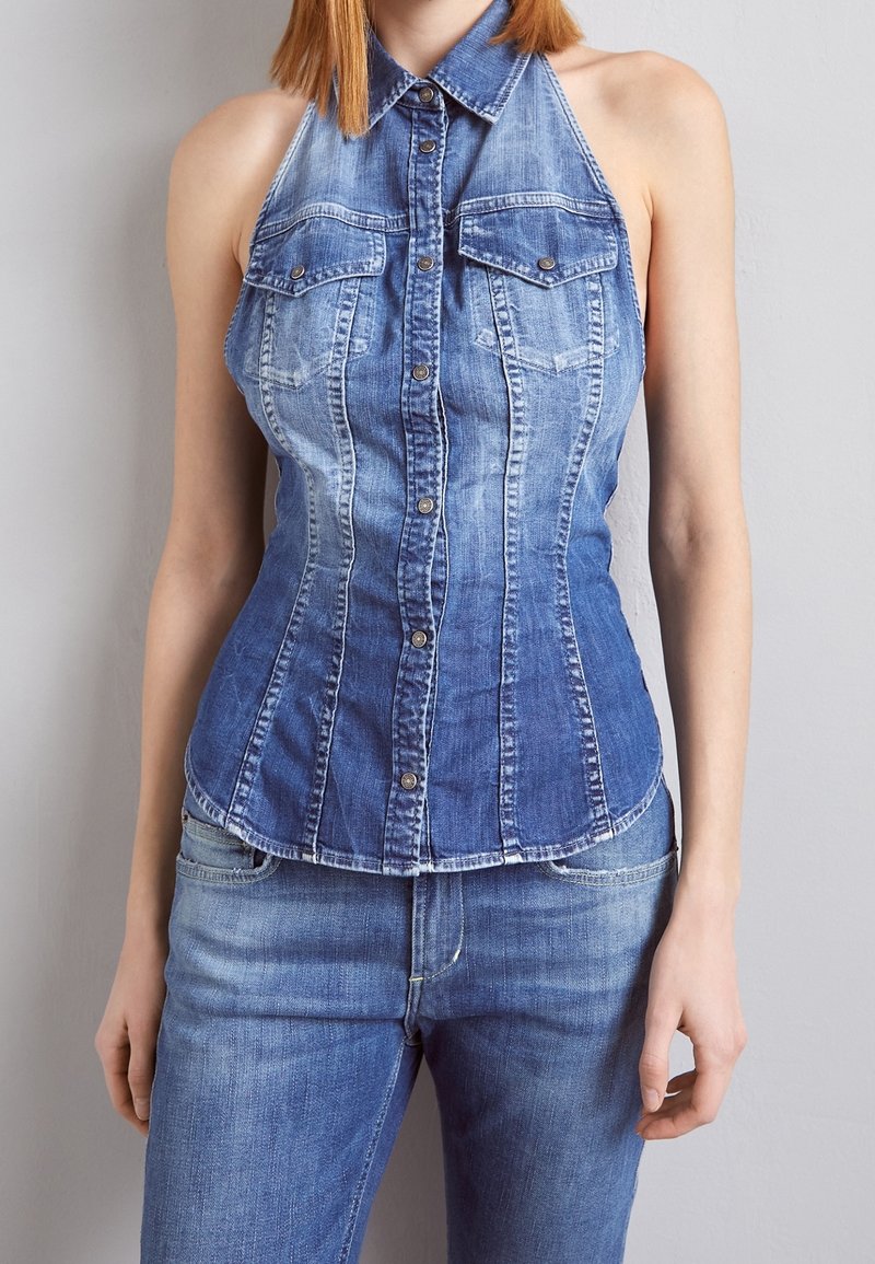 Femme portant un haut en jean bleu sans manches avec col et poches devant, assorti à un jean bleu sur fond gris.