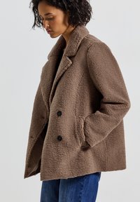 Veste en polaire marron avec une coupe décontractée, col à revers et deux boutons noirs. Comprend des poches latérales et une surface douce et texturée.