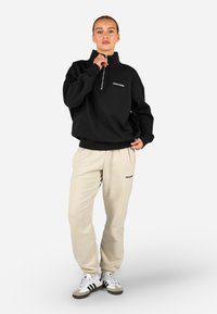 Melns ceturtdaļas rāvējslēdzēja džemperis ar logo, apvienots ar gaiši brūnām treniņbiksēm, kurām ir logo. Baltas sporta apavi ar melnām un pelēkām akcentiem.