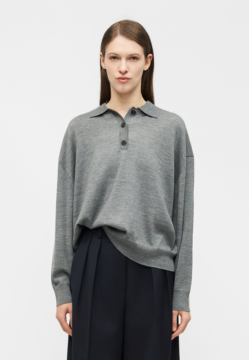 Γυναίκα με ίσια καστανή τρίχα που φοράει χαλαρό γκρι πλεκτό πόλο με μακριά μανίκια και μαύρο ψηλόμεσο παντελόνι με πιέτες, στέκεται μπροστά σε μια μονόχρωμη επιφάνεια.