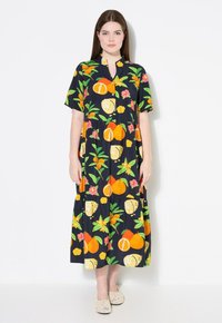 Vestido negro con un estampado colorido de frutas y flores, mangas cortas, cierre de botones en la parte frontal y un diseño de falda en capas. Llevado con zapatos beige.