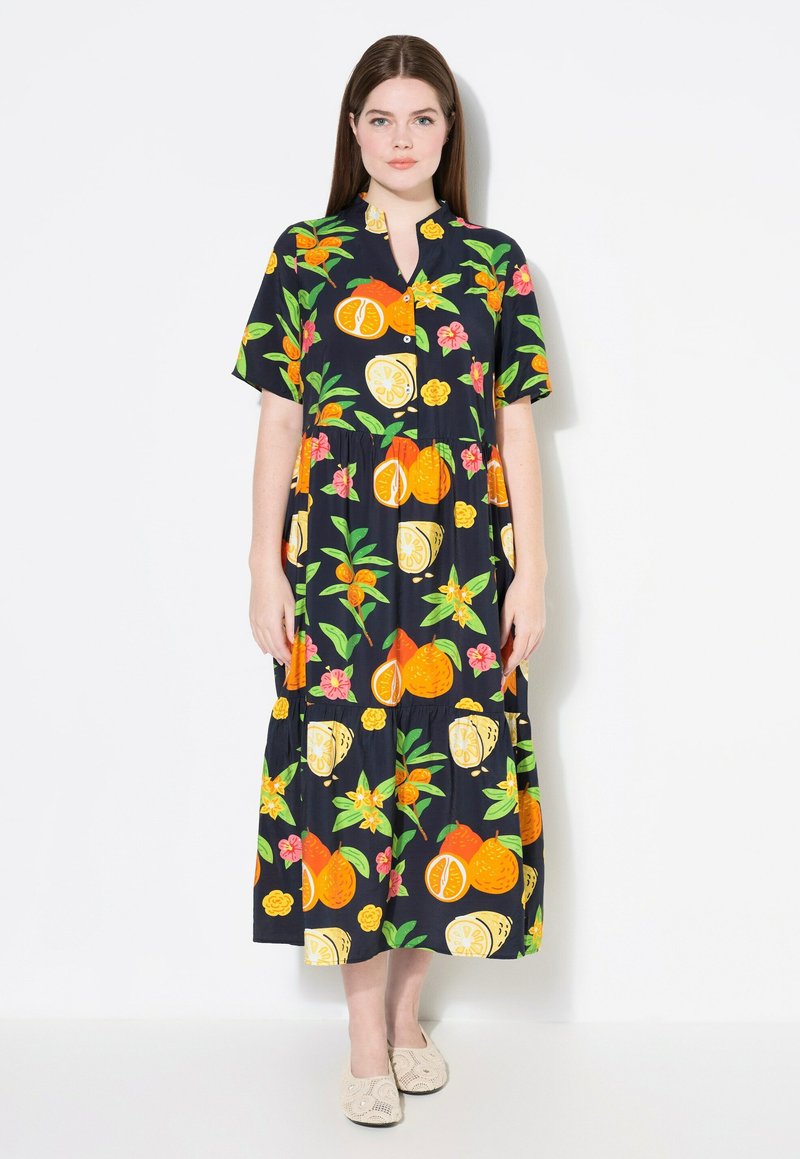 Vestido negro con un estampado colorido de frutas y flores, mangas cortas, cierre de botones en la parte frontal y un diseño de falda en capas. Llevado con zapatos beige.