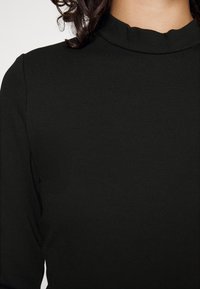 Buste d'une personne portant un haut à manches longues noir avec un col arrondi légèrement froncé, et des cheveux bouclés foncés visibles.