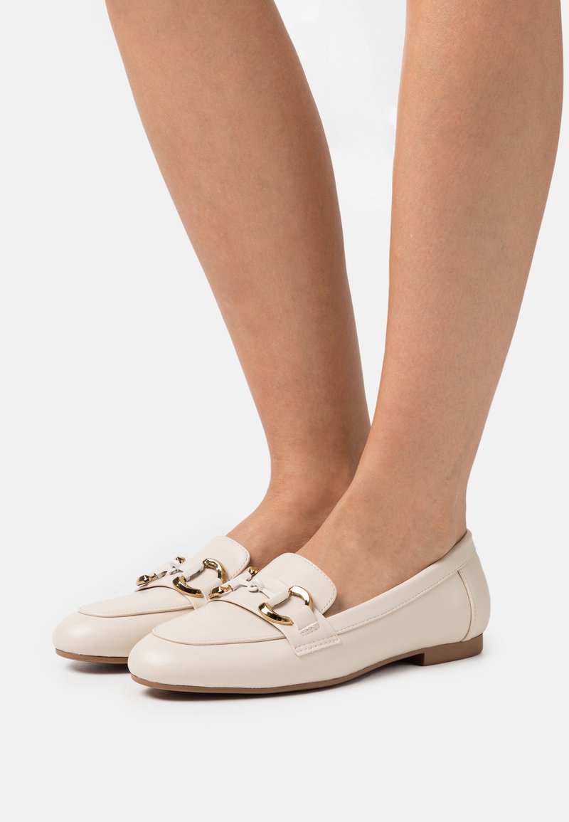 Pieds portant des mocassins en cuir crème avec un détail mors en or, debout sur une surface blanche unie.