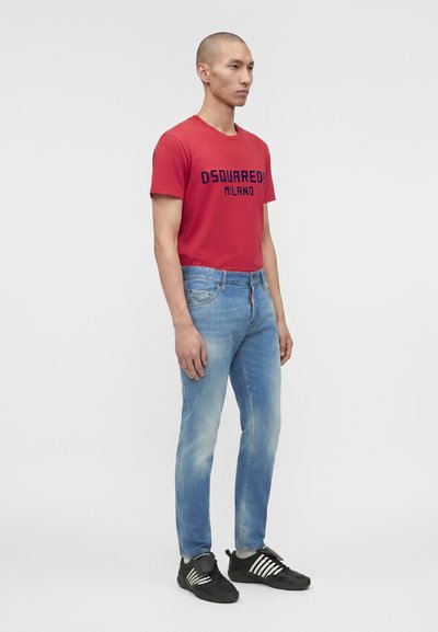 Άνδρας φοράει κόκκινο t-shirt "DSQUARED2 MILANO", ανοιχτό μπλε τζιν και μαύρα αθλητικά παπούτσια με λευκές ρίγες, στέκεται μπροστά σε απλό φόντο.