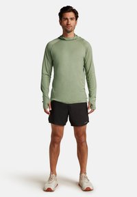 Maglietta a maniche lunghe verde chiaro con cappuccio, abbinata a pantaloni corti neri. Il modello indossa scarpe da ginnastica beige. Tessuto liscio, vestibilità atletica e design semplice.