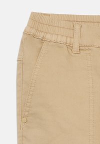 Pantalons en coton beige avec une taille élastique, des poches avant et des détails de couture subtils. Le tissu a une texture lisse et mate.