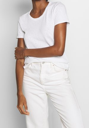 Eine Person trägt ein einfaches weißes T-Shirt mit kurzen Ärmeln und hochgeschnittene weiße Jeans, die vor einem schlichten hellgrauen Hintergrund steht.