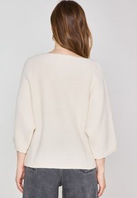 Pull tricot écru avec des manches larges ¾, tissu texturé, ourlet brut et coupe décontractée. L'arrière-plan est neutre.