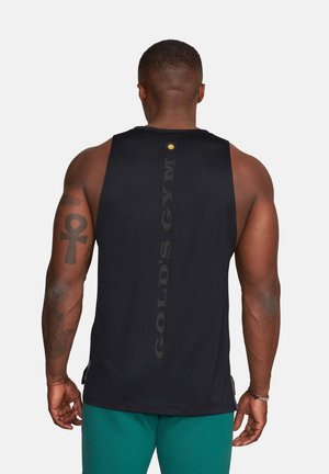 Mann mit kurzen Haaren, der ein schwarzes, ärmelloses Gold’s Gym-Shirt und türkise Shorts trägt, Tattoos auf seinem linken Arm zeigt und von hinten zu sehen ist.