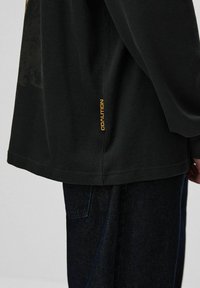 Czarna, teksturowana koszula z długim rękawem, z bocznym rozcięciem i żółtą metką "COALITION" na dole, zestawiona z ciemnymi dżinsami.