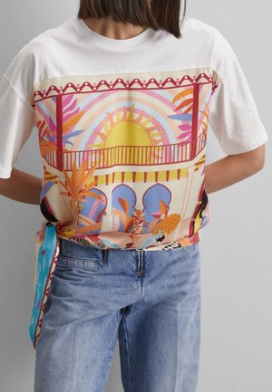 Kobieta w białej koszulce z kolorowym abstrakcyjnym nadrukiem słońca, roślin i balkonu, zestawiona z jasnoniebieskimi dżinsami.