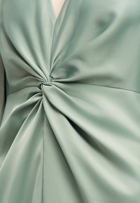 Detailný záber na šalviovozelenú látku stočenú a zhromaždenú v strede, vytvárajúcu záhyby a textúru na hladkom materiáli.