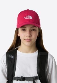 Ljusrosa basebollkeps med en böjd skärm, med vit broderad logotyp framtill. Svarta remmar från en ryggsäck är synliga.