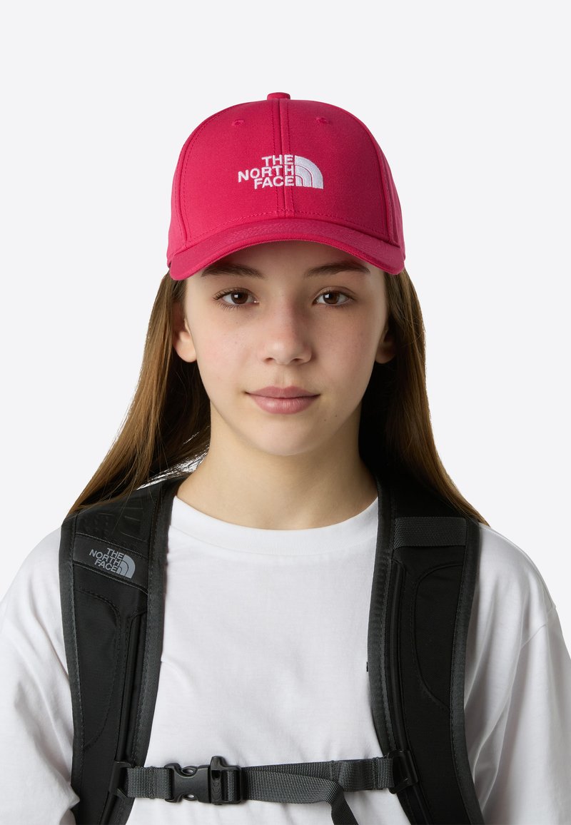 Ljusrosa basebollkeps med en böjd skärm, med vit broderad logotyp framtill. Svarta remmar från en ryggsäck är synliga.