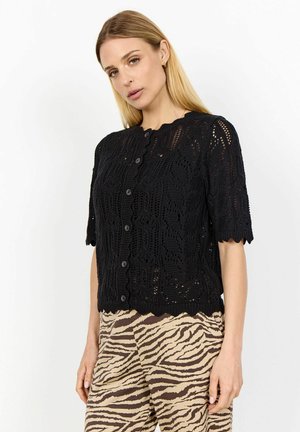 Zwarte gebreide cardigan met korte mouwen, gegolfde zoom, textuur van kant en zes knopen aan de voorkant. Gecombineerd met gestreepte beige broek.