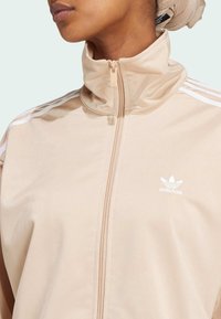 Béžová sportovní bunda s vysokým límcem, která má bílé pruhy na ramenou a bílé logo. Hladká, lehká textilie.