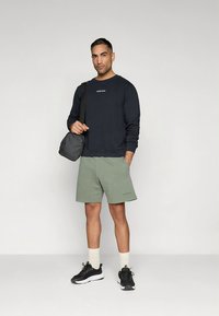 Sweatshirt azul-marinho com logótipo e calções verde-oliva. Ténis desportivos pretos com solas brancas e meias creme até à canela. Pequena mala cinza ao ombro.