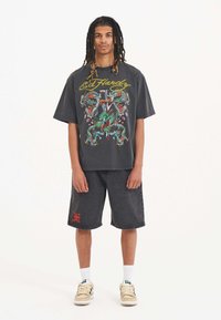 A young individual stands confidently, wearing a black oversized Ed Hardy t-shirt with colorful dragon graphics and gray denim shorts.