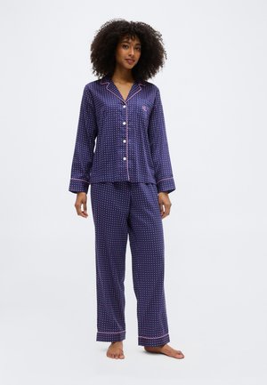 Vrouw met krullend haar die een marineblauw zijden pyjama met roze polkadots en bies draagt, blootsvoets staand tegen een effen achtergrond.