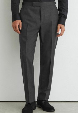 Man met donkergrijze formele broek met verstelbare lussen aan de zijkant, zwart overhemd en zwarte suède schoenen, staand tegen een lichte muur.