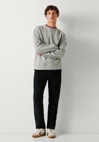 Grijze sweatshirt met rode halslijn, uitgerust met een klein "PJL" logo. Gecombineerd met donkere broek en beige/witte sneakers. Glad textuur.