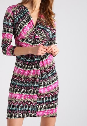 Mujer que lleva un vestido de manga larga hasta la rodilla con un patrón geométrico multicolor en rosa, negro, azul verdoso y beige.