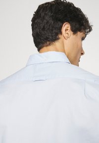 Homme aux cheveux courts, bouclés et foncés, portant une chemise bleu clair à col, vu de dos et légèrement tourné sur le côté.