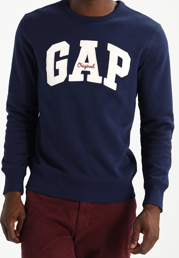Homme portant un sweat-shirt bleu marine avec les lettres blanches "GAP" et un petit texte rouge "Original", associé à un pantalon rouge foncé.