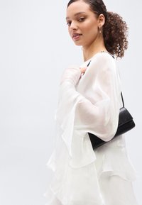 Νεαρή γυναίκα με σγουρά μαλλιά, φορώντας λευκό μακρυμάνικο πουκάμισο με βολάν, κρατά μια μικρή μαύρη τσάντα ώμου σε ανοιχτό φόντο.