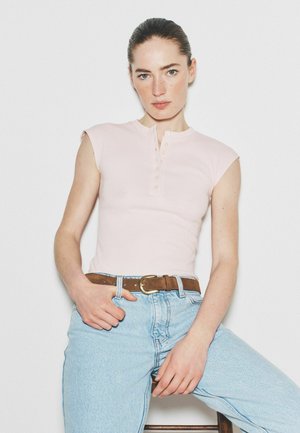 Kvinde med mørkt hår trukket tilbage iført ærmeløs, lyserød knappet top, lyseblå højtaljede jeans og brun bælte, siddende på en skammel.