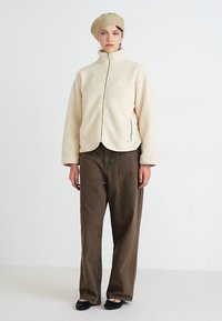 Cremfarbener Fleece-Reißverschlussjacke mit hohem Kragen, kombiniert mit braunen weiten Hosen. Das Model trägt außerdem eine strukturierte beigefarbene Strickmütze.