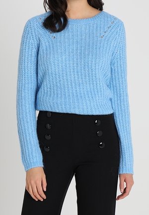 Pull en tricot bleu clair avec un motif texturé, col rond, et petites découpes au niveau des épaules, associé à un pantalon noir avec des boutons.
