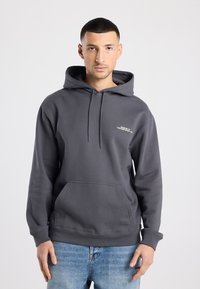 Donkergrijze hoodie met een voorvak, capuchon met trekkoord en een wit tekstlogo. Het materiaal lijkt zacht en licht dik. Aansluitende boorden en zoom.