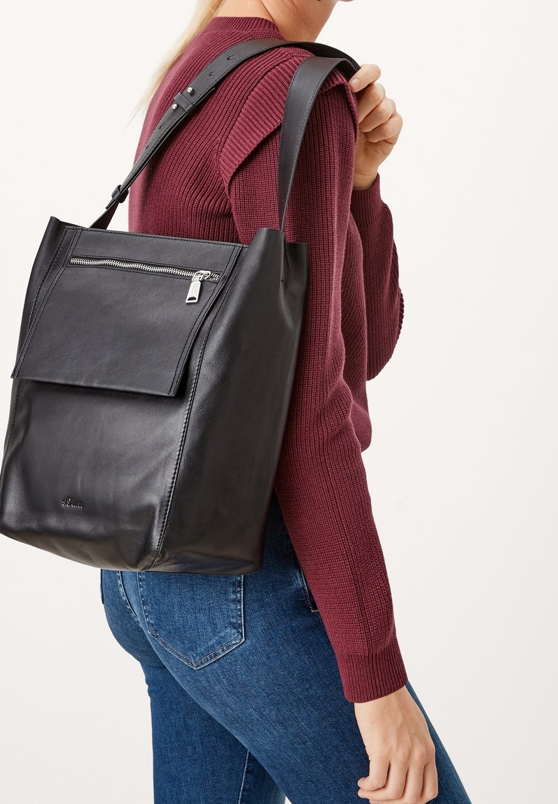 Femme portant un pull bordeaux et un jean bleu, portant un grand sac à bandoulière en cuir noir avec une poche zippée à l'avant.