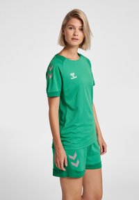 Grön sporttröja och shorts-set, tillverkat av andningsbart meshmaterial. Har grå chevrondetaljer på axlarna och shortsen.