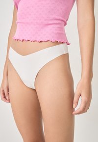 Tanga de algodón blanco con una cintura baja y una ligera forma de V, combinada con una blusa rosa con un patrón texturizado de corazones y un dobladillo con forma de olas.