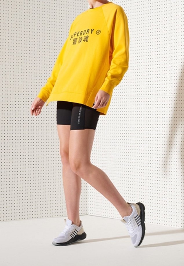 Gul sweatshirt med svart logotyp, svarta shorts och vita sportskor med svarta accenter; person står på en neutral bakgrund.