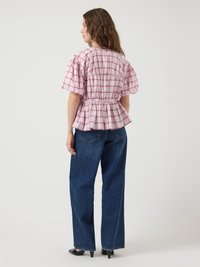Blusa a scacchi rosa e bianca con maniche a palloncino, vita arricciata e orlo peplo, abbinata a jeans blu a vita alta e tacchi neri.