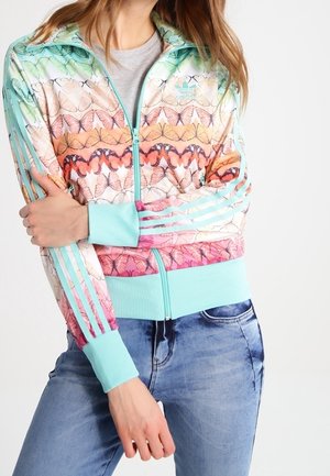 Femme portant une veste zippée à motif papillon pastel avec des poignets et des bandes turquoise, associée à un jean bleu.