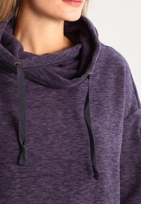 Felpa viola con un grande collo a cowl, cordini neri e un tessuto morbido e texture. I dettagli includono accenti dorati al collo.