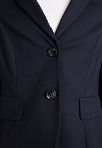 Marineblå blazer med en strukturert krage, tre knapper og to framlommer. Stoffet har en glatt tekstur og lett stretch.