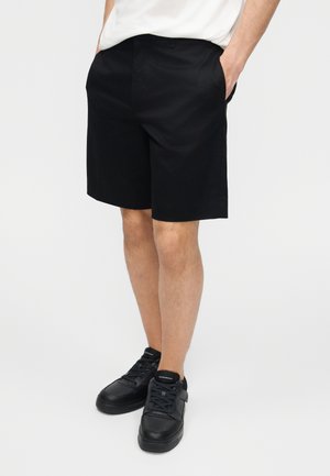 Vyras dėvi juodus iki kelių ilgio šortus, baltą marškinėlį ir juodus sportbačius, stovintis rankas kišenėse ant vienspalvio fono.