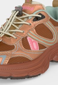 Gros plan d'une basket marron et pêche avec un logo de marque rose, des lacets élastiques blancs avec un verrou gris, une bordure bleu clair et une semelle en caoutchouc texturée.