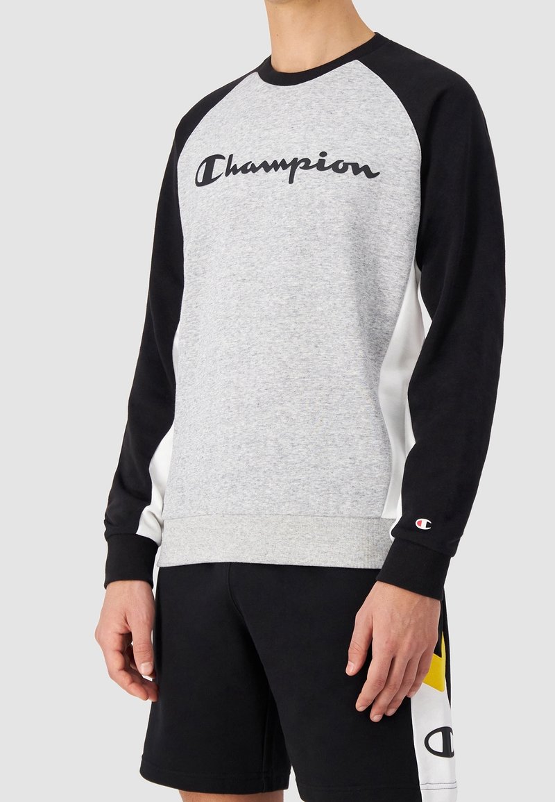 Personne portant un sweat-shirt gris et noir Champion avec logo sur la poitrine et le poignet gauche, associé à un short noir avec des détails blancs et jaunes.